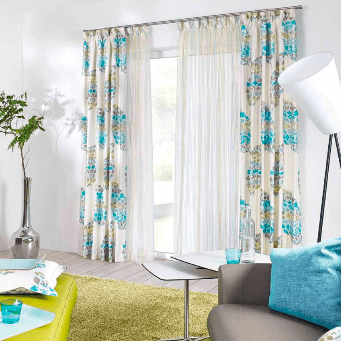 Modern living room with floral curtains, colorful cushions, and a sleek lamp beside a large window.